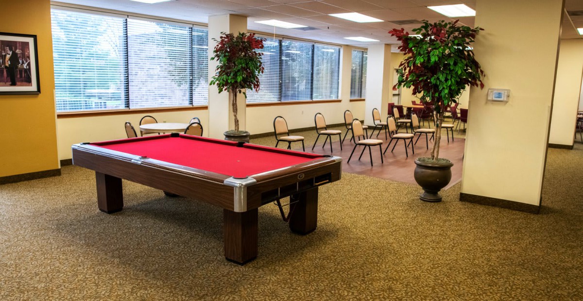A spacious recreation room featuring a red pool table, chairs arranged along the walls, large windows, and potted plants on a carpeted floor.