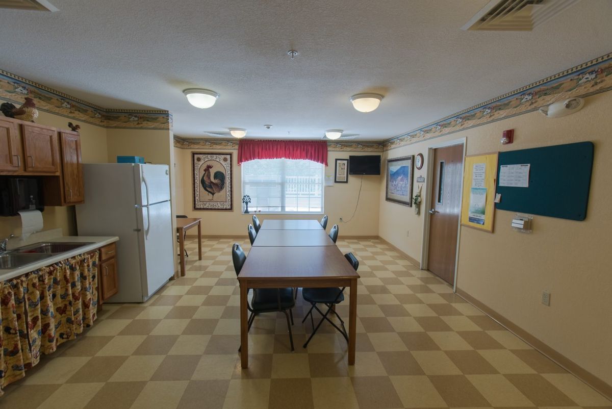 A communal dining area with a checkered floor, featuring a table and chairs, refrigerator, window with curtains, and various artworks on the walls.