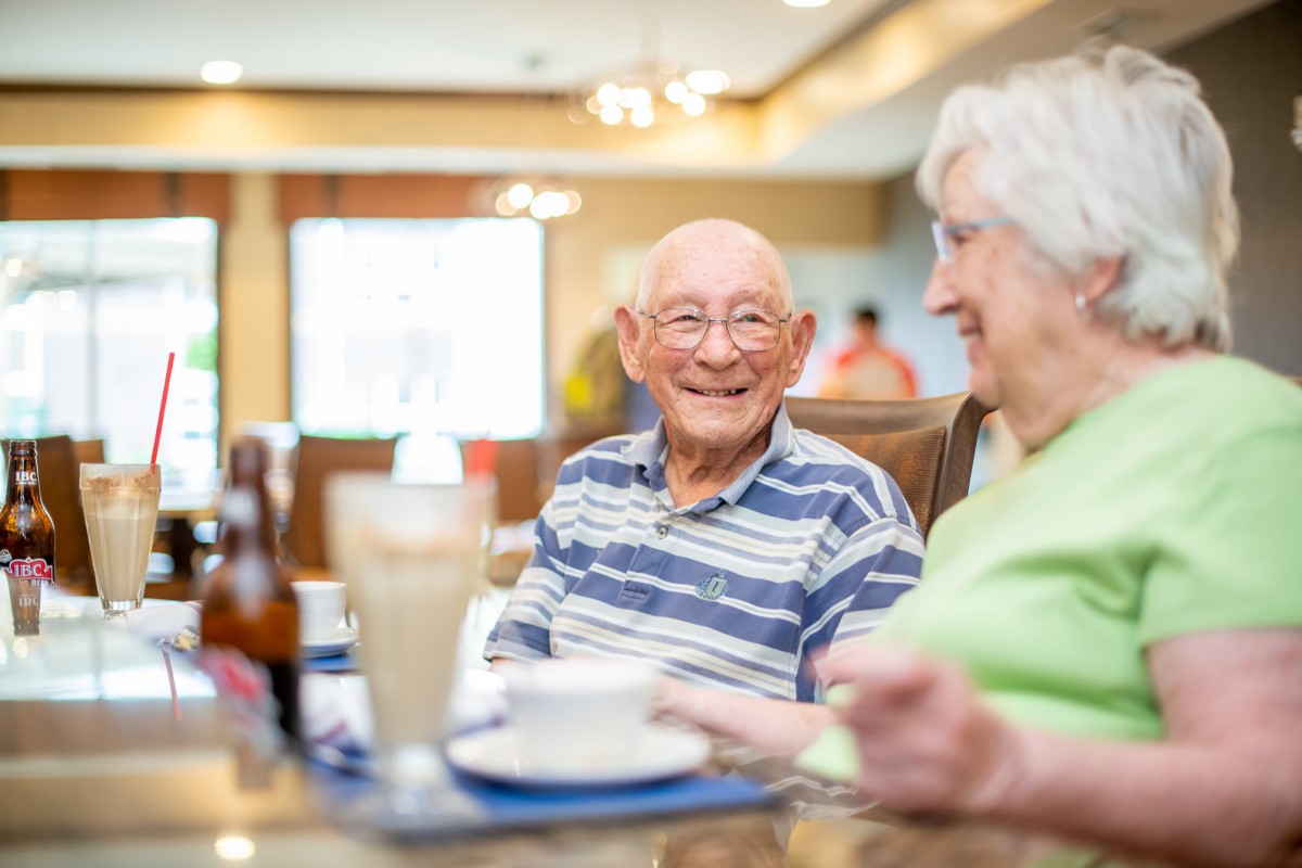Two senior individuals enjoying a conversation over a meal in a bright dining area.