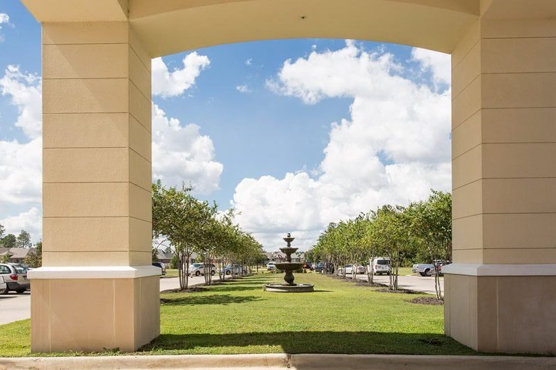 A scenic view through an archway featuring a fountain surrounded by trees and a partly cloudy sky.