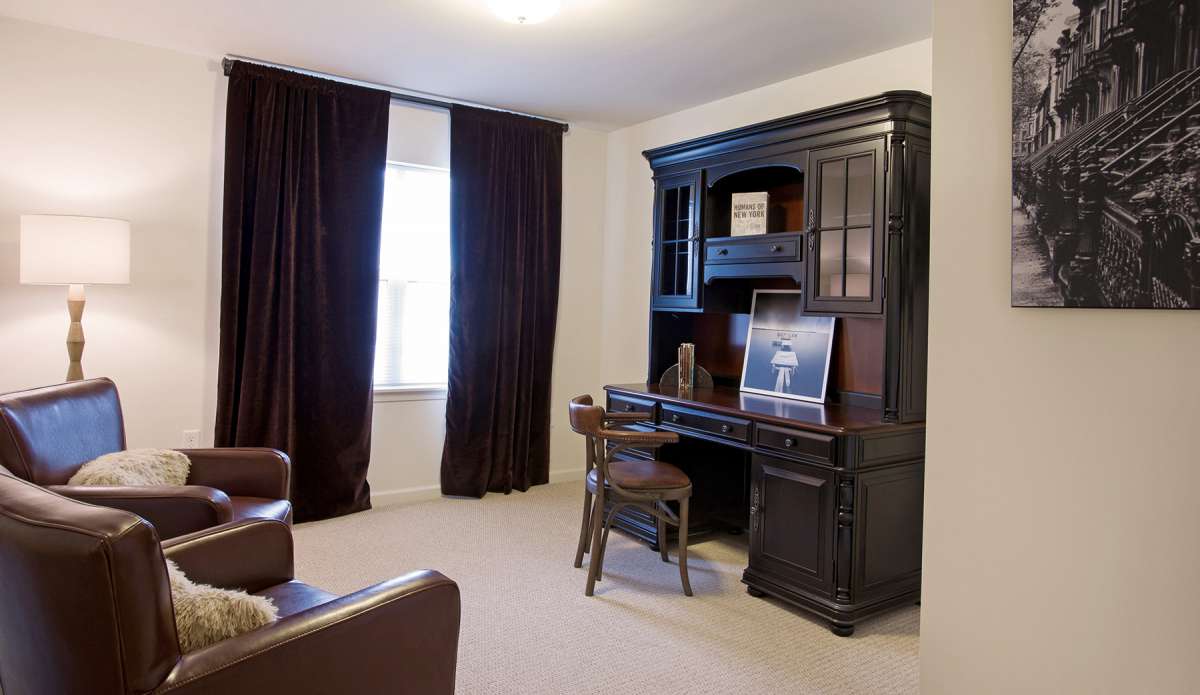 A home office with a dark wooden desk and bookshelf, accompanied by a chair and window with curtains.