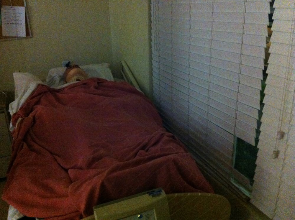 A hospital room with a patient lying in bed covered by a blanket, next to a window with blinds.