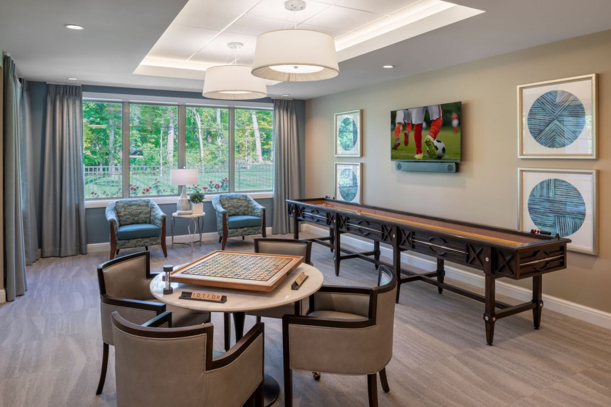 A modern game room featuring a card table, shuffleboard, and comfortable seating with large windows providing natural light.