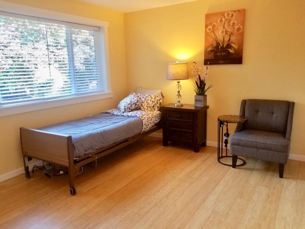 A cozy bedroom with a single bed, nightstand, lamp, and chair on a wooden floor.