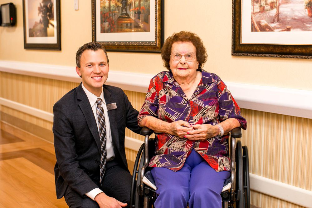 A man in a suit kneels beside an elderly woman sitting in a wheelchair, both smiling at the camera indoors.