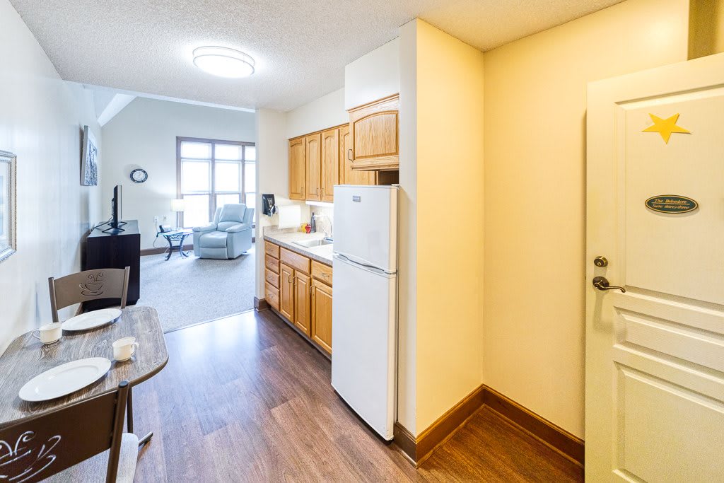 A cozy apartment interior featuring a compact kitchen with wooden cabinets and a fridge, leading into a bright living area.
