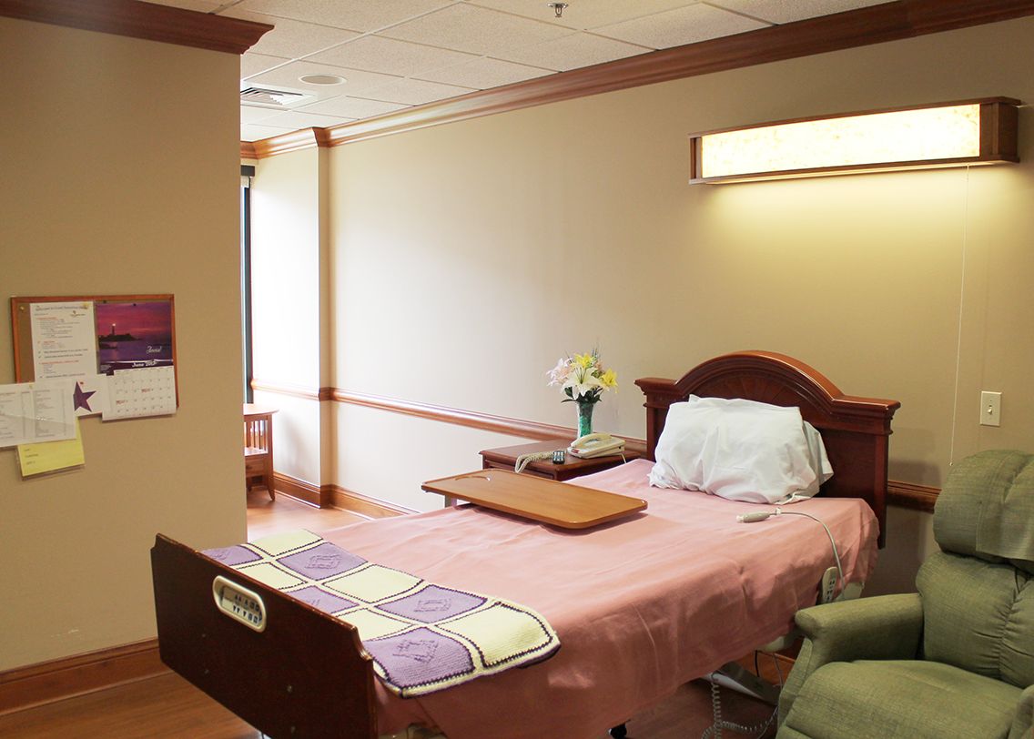 A hospital room featuring a patient bed with pink sheets, a side table, and a chair.