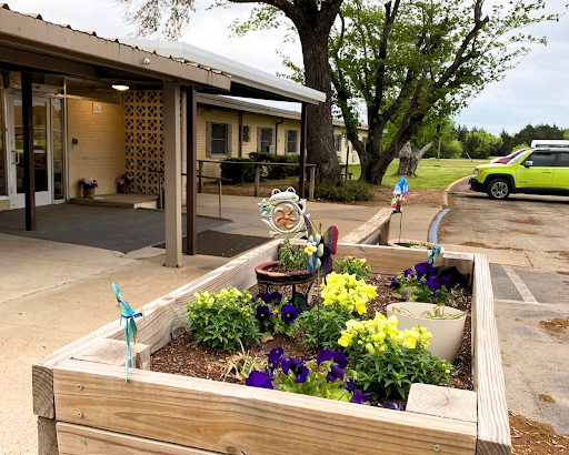 A vibrant garden with yellow and purple flowers planted in a wooden box, set against a backdrop of a building and trees.