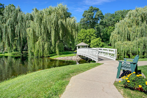 A serene park scene featuring a bridge over a calm body of water, surrounded by lush greenery and willow trees.