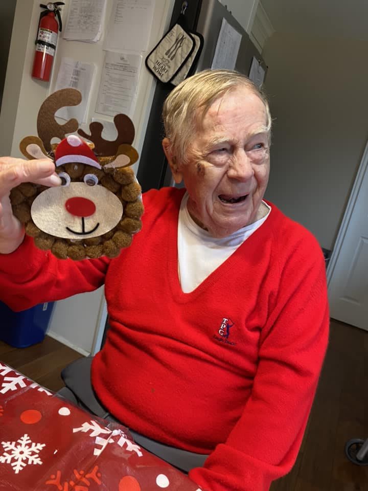 An elderly man wearing a red sweater is smiling and holding a festive reindeer decoration.