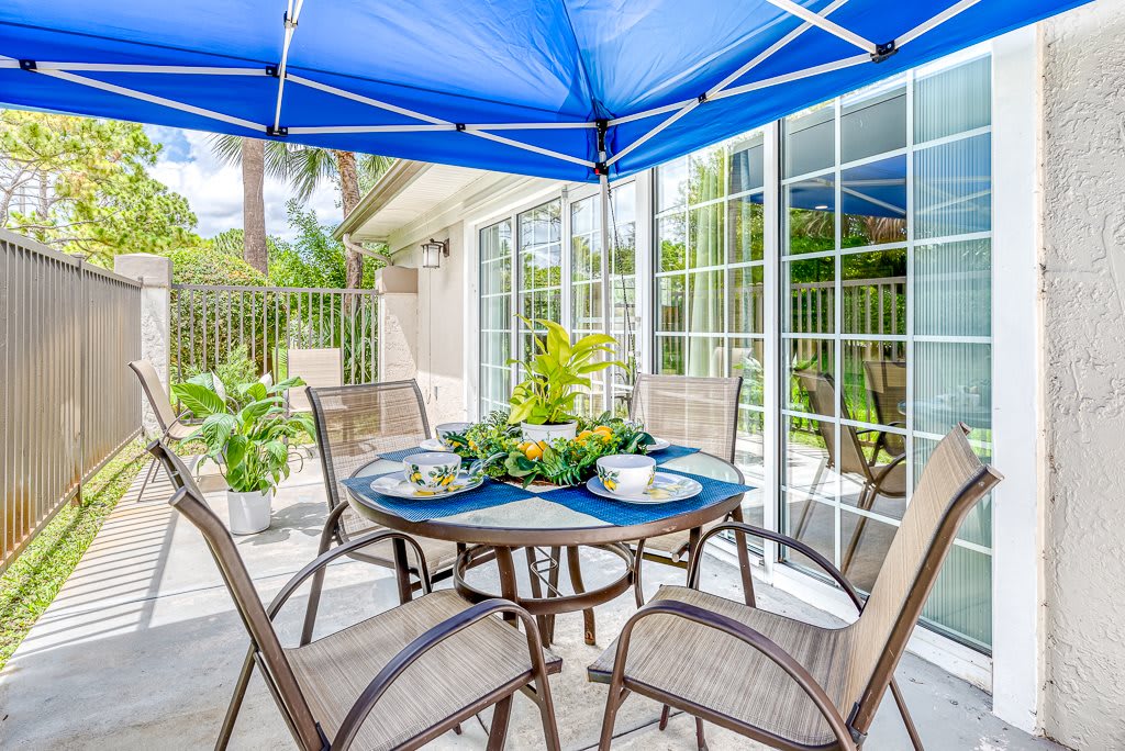 Outdoor patio with a table set under a blue canopy, surrounded by chairs and greenery.