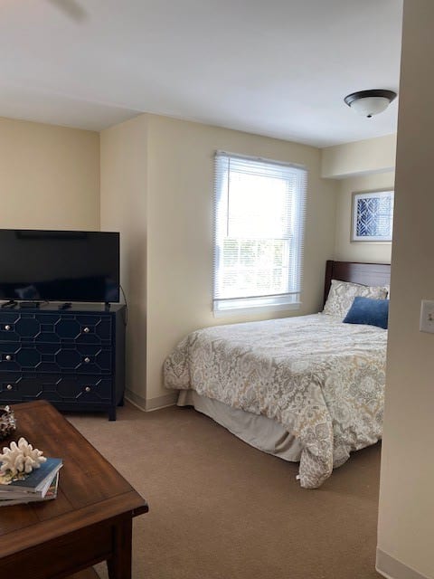 A cozy bedroom featuring a bed with patterned bedding, a dresser with a television on top, and a window allowing natural light to brighten the room.