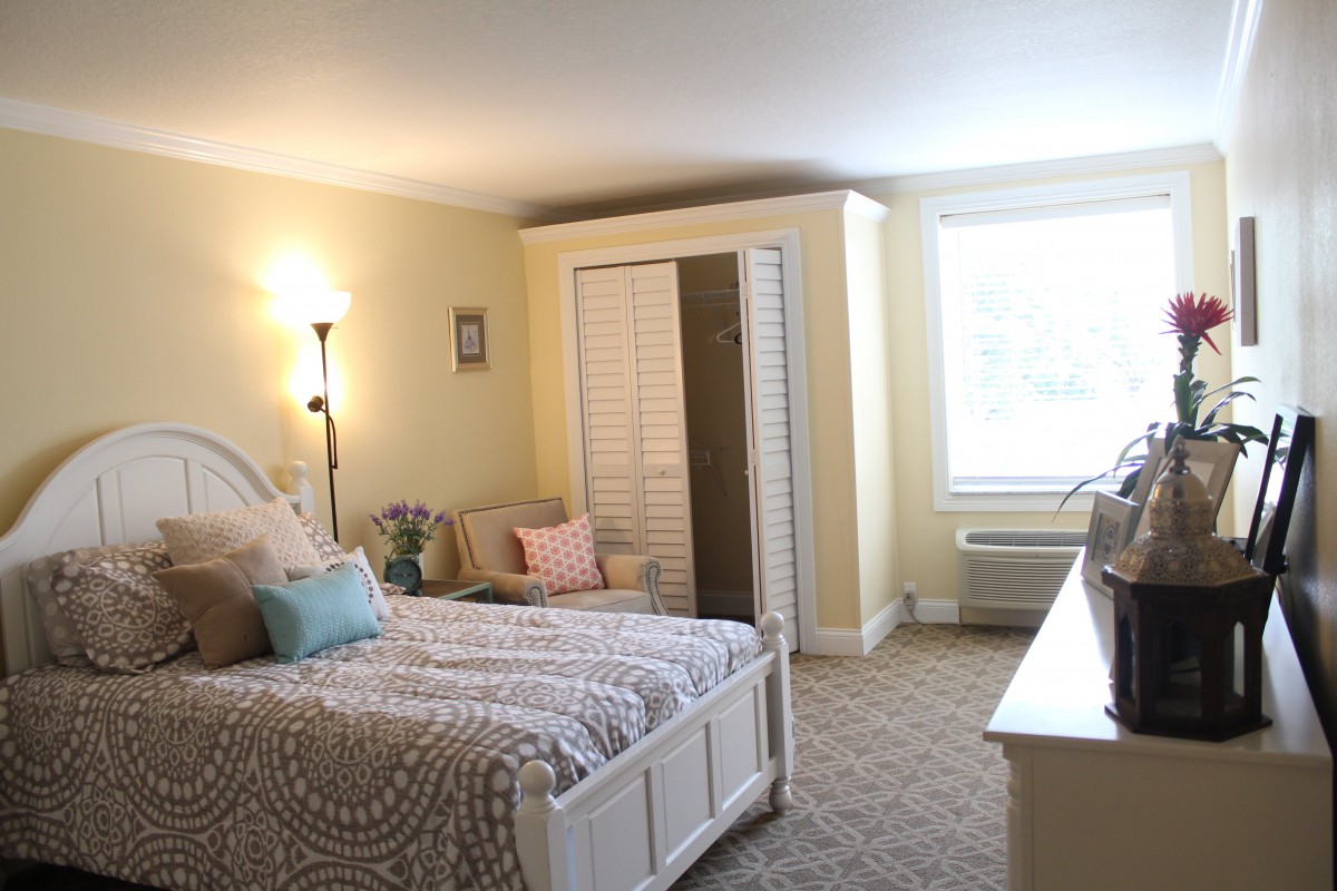 A cozy and well-lit bedroom featuring a bed, floor lamp, and a wardrobe with an open closet door. The room has light-colored walls and carpeted flooring.