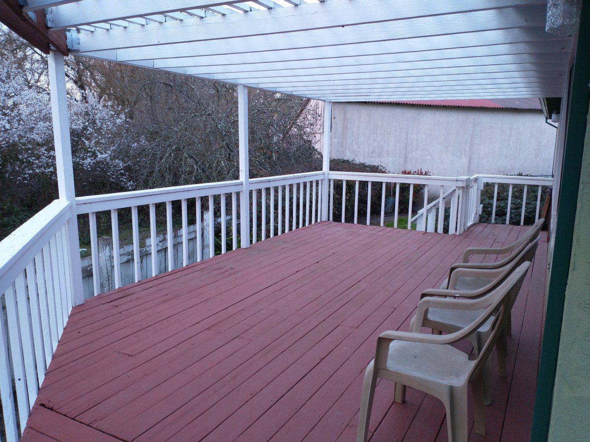 A spacious wooden deck with a pergola overhead and several plastic chairs arranged along the side.