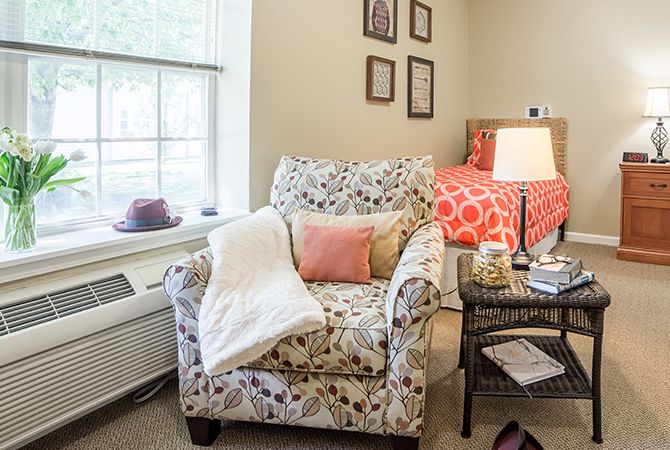 A cozy bedroom with a floral armchair, side table with a lamp, and a bed with colorful bedding.