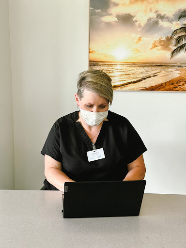 A woman in professional attire working on a laptop while wearing a mask in an office setting.