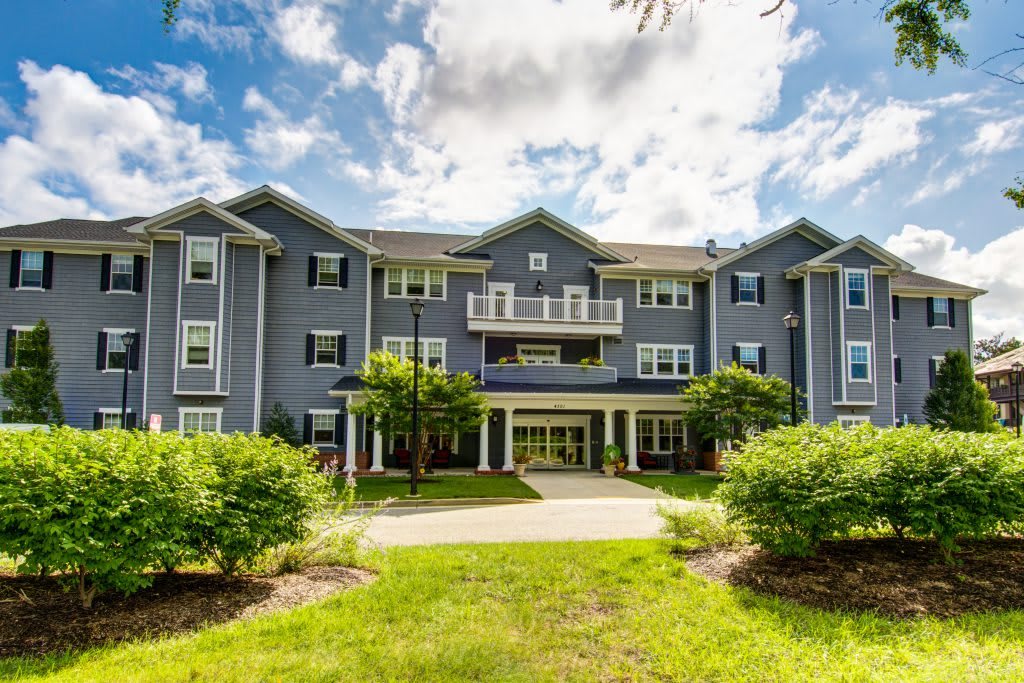 A modern residential apartment building with a landscaped front yard under a partly cloudy sky.