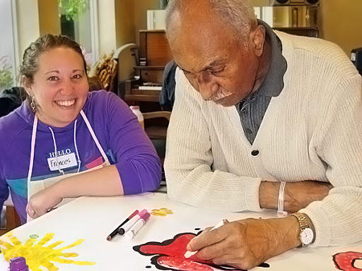 A joyful art session where an elderly man is engaged in painting, assisted by a smiling caregiver.