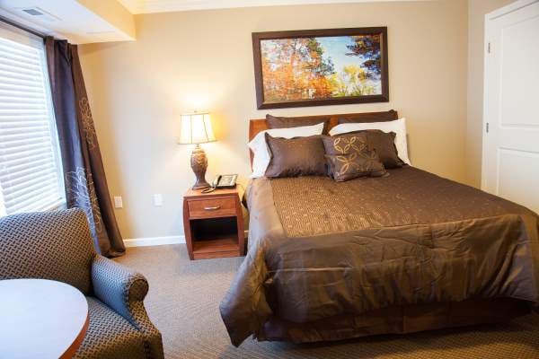 A cozy bedroom with a large bed, bedside table, and lamp, decorated with warm tones and a nature painting on the wall.