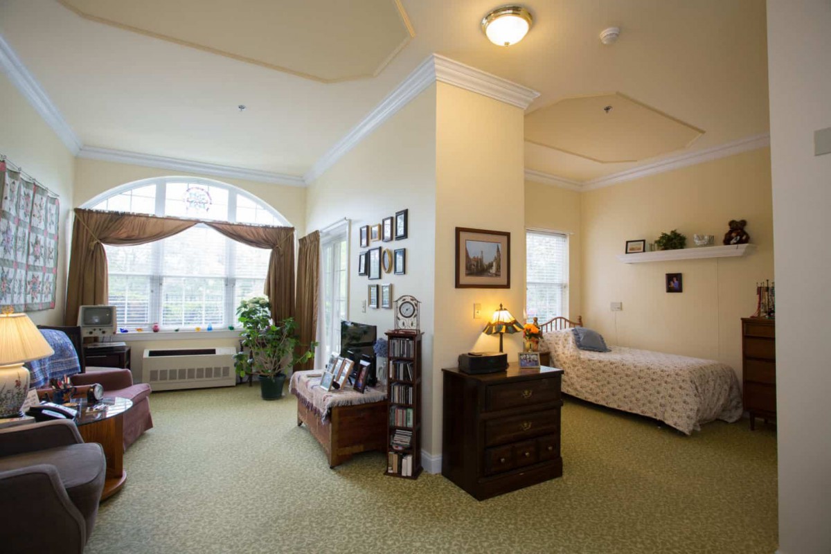 A cozy bedroom with warm lighting, featuring a bed, dresser, and a large window letting in natural light.