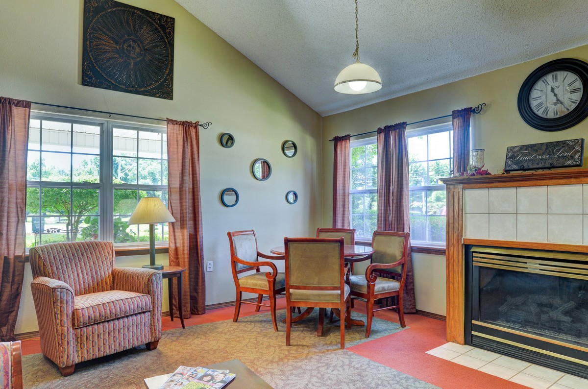 A cozy living room with a small dining area, featuring stylish furniture, large windows, a fireplace, and decorative wall elements.