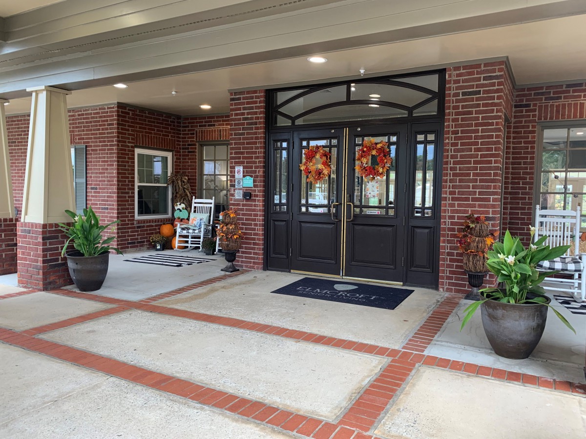 Entrance of a building with double doors, brick facade, potted plants, and outdoor seating.