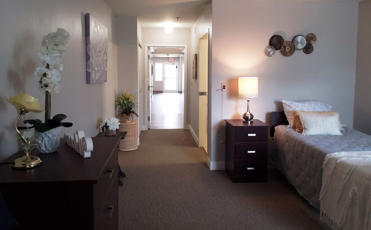 A cozy bedroom with a hallway view, featuring a bedside table and lamp.