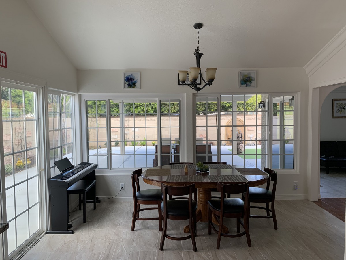 A bright dining room with a table, chairs, and a piano, illuminated by natural light through large windows.