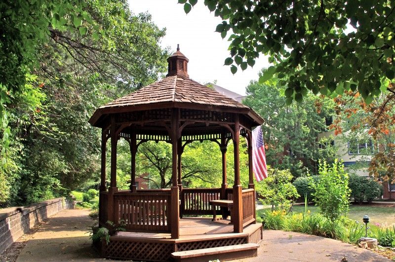 A wooden gazebo in a lush garden setting, surrounded by greenery.