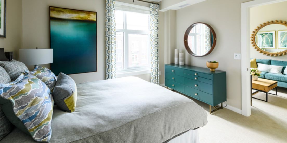A modern bedroom featuring a comfortable bed, large window, abstract artwork, and a stylish dresser with a round mirror.