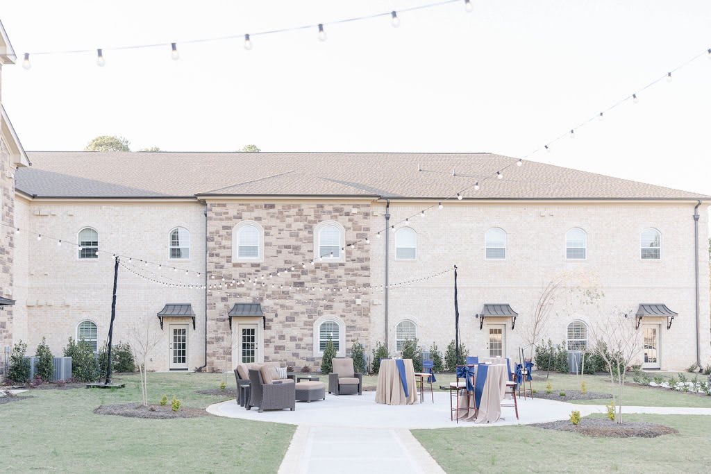 A building with a stone facade features outdoor seating on a patio, adorned with string lights, and surrounded by a well-maintained lawn.