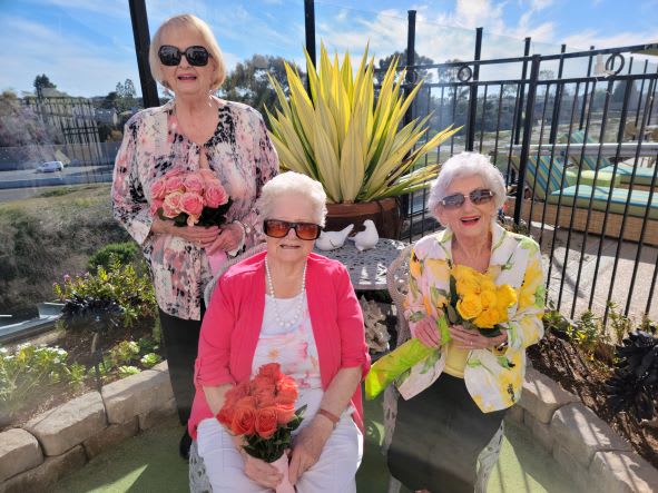 Three women wearing sunglasses hold colorful flowers while sitting in a sunny garden.
