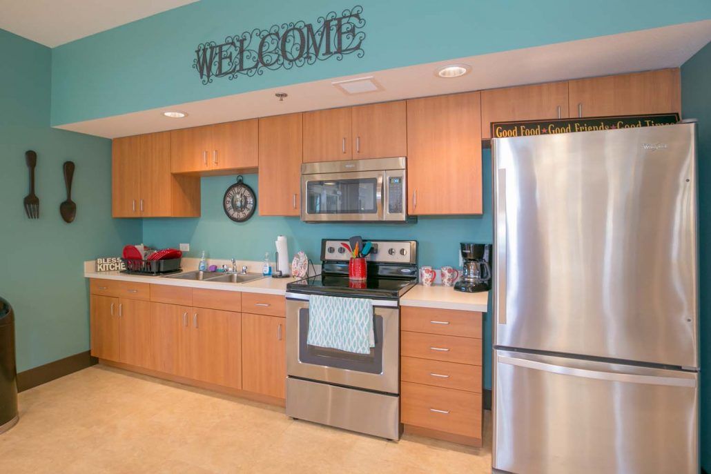 A modern kitchen with stainless steel appliances and wooden cabinets, featuring a welcome sign on blue walls.