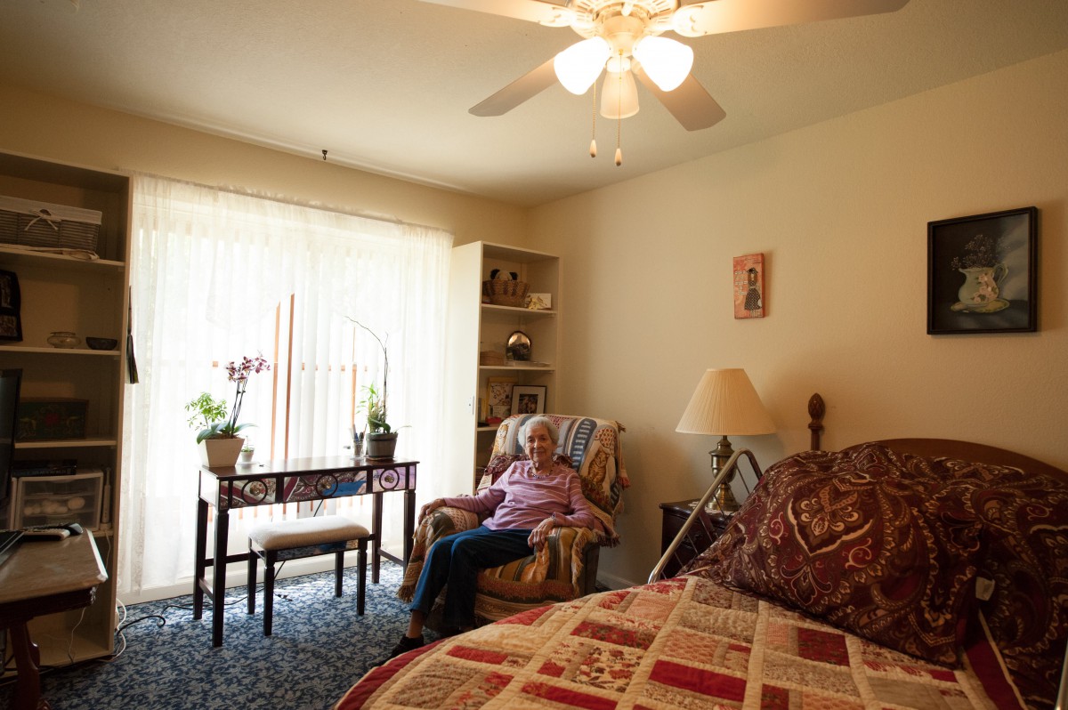 An elderly person sitting in a cozy bedroom with natural light streaming in through the window.