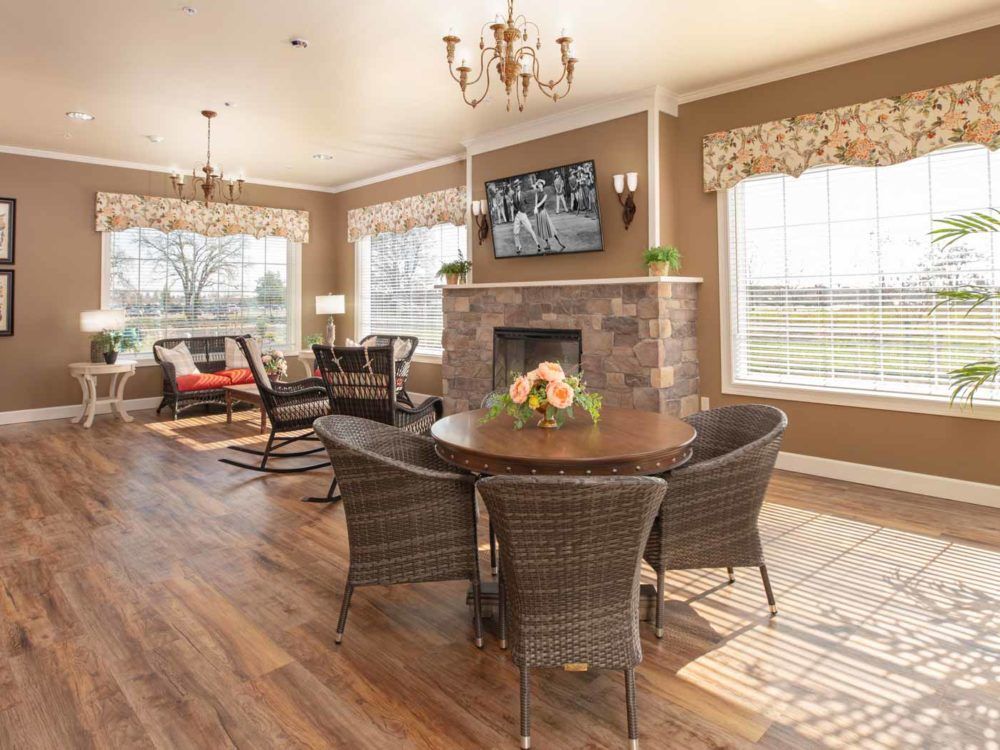 A spacious and elegantly furnished living room with a fireplace and large windows, featuring a round dining table and wicker chairs.