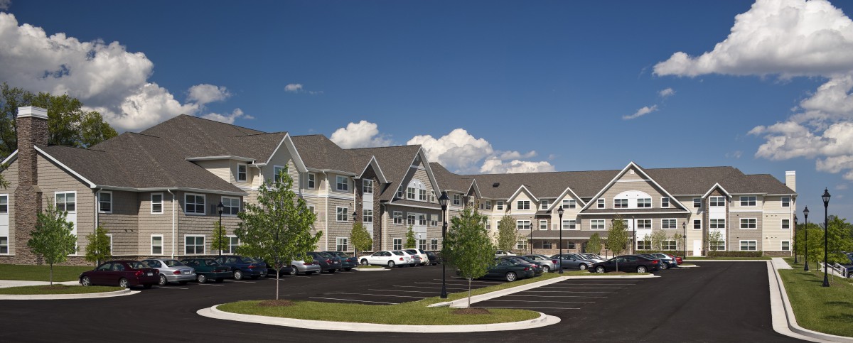 A modern residential apartment complex with a parking lot and trees under a clear blue sky.