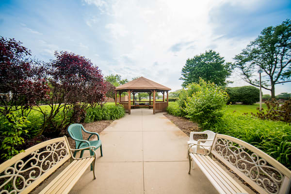 A peaceful garden with a pathway leading to a wooden gazebo, surrounded by lush greenery and benches.