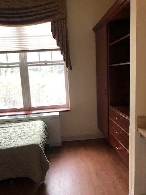 A simple bedroom with a bed, large window, and wooden wardrobe.