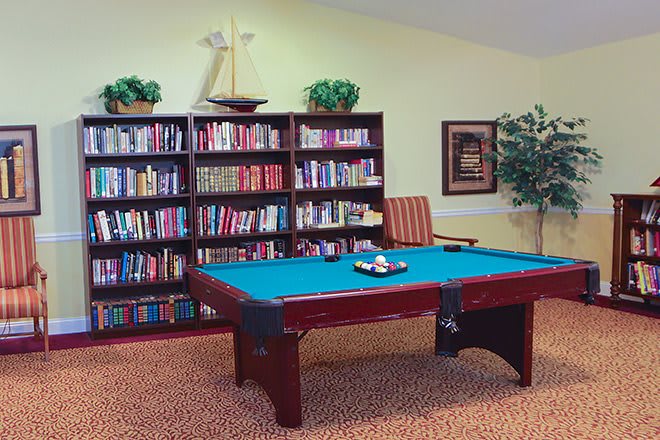 A cozy room featuring a billiard table surrounded by bookshelves filled with books, decorated with plants and a model sailboat.
