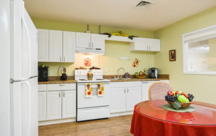 A cozy kitchen with white cabinets, a stove, and a dining area featuring a fruit bowl on a red tablecloth.