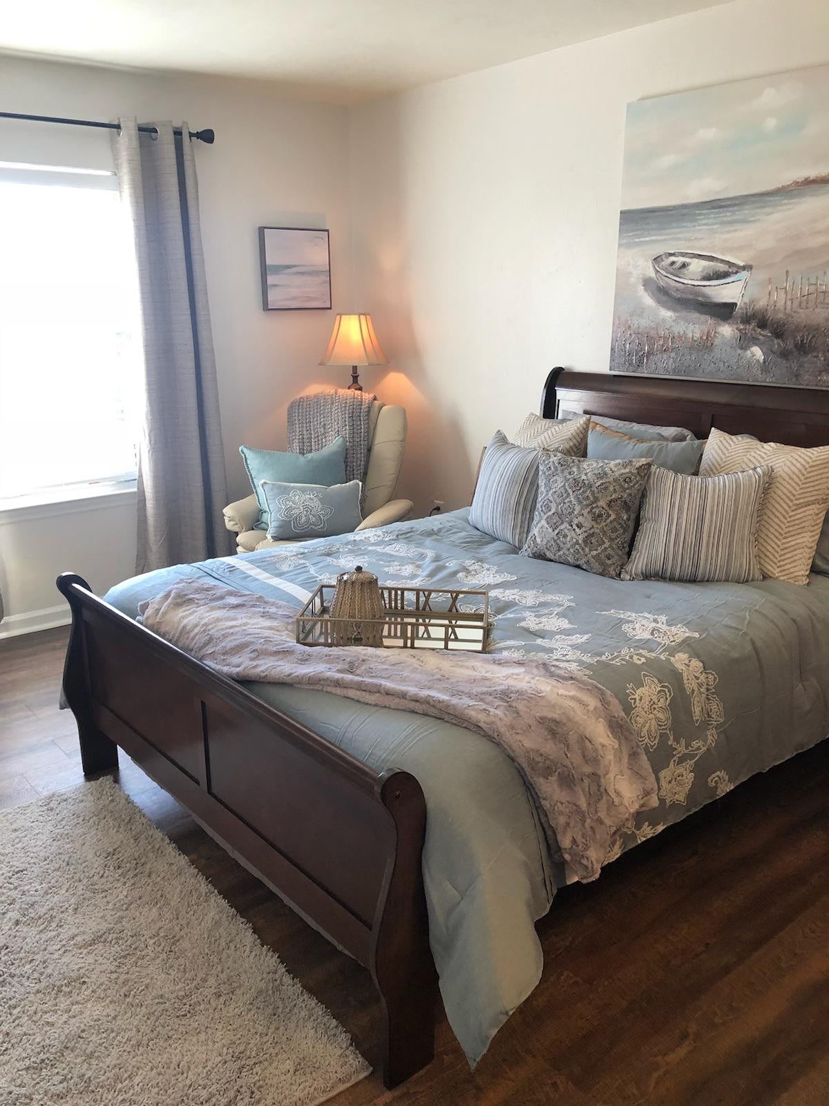 A cozy bedroom with a wooden bed frame, stylish bedding, and a reading nook with an armchair and lamp.