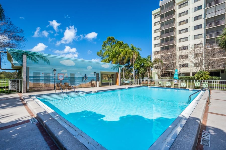 A luxurious outdoor swimming pool surrounded by palm trees and a high-rise building, set against a sunny tropical backdrop.