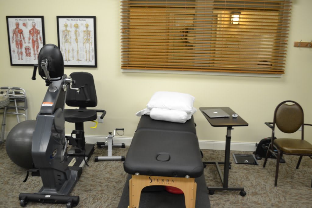 A therapy room equipped with a treatment table, exercise machine, and anatomical poster.