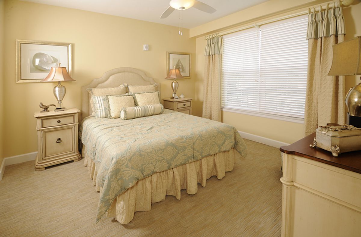 A cozy bedroom with a neatly made bed, nightstands, lamps, and a large window with blinds and curtains.