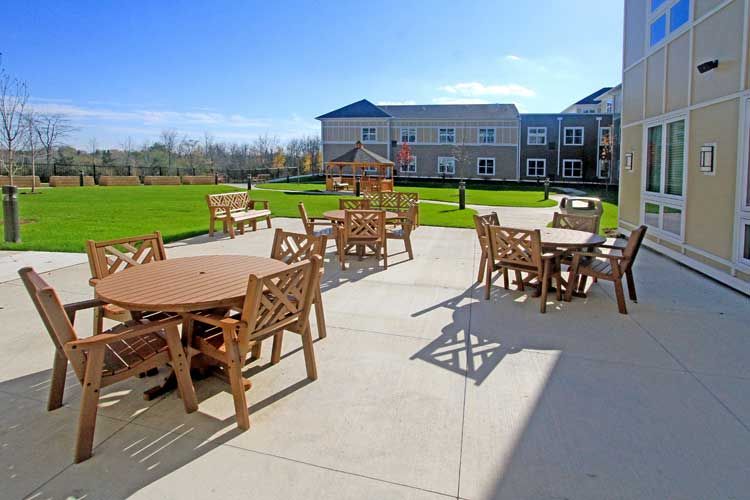 A sunny patio area with tables and chairs overlooking a well-maintained lawn and building.