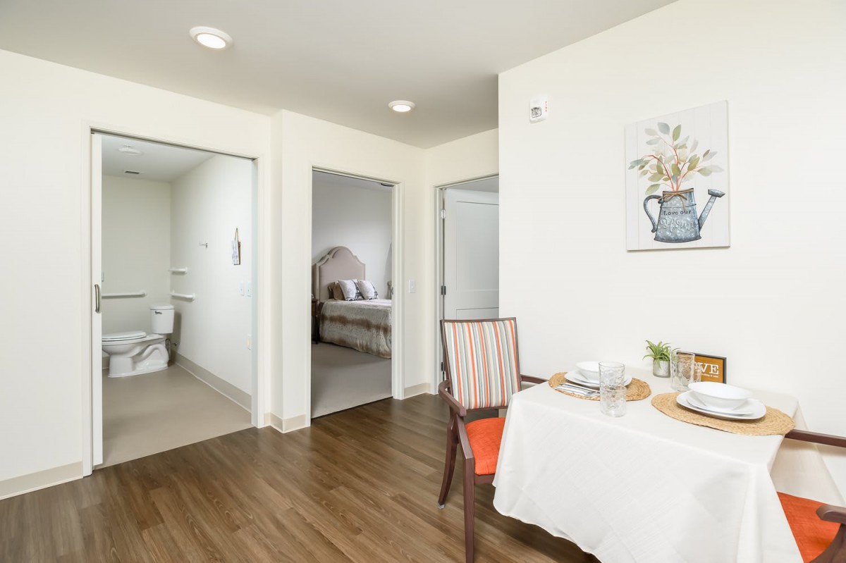 A modern apartment interior featuring a dining area with a set table, and views into a bedroom and bathroom.