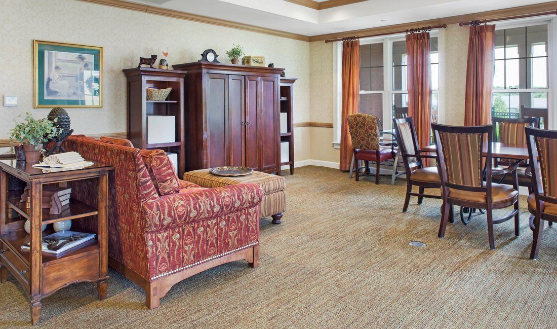 A cozy living room with a patterned armchair, wooden cabinet, and dining table set near a window.