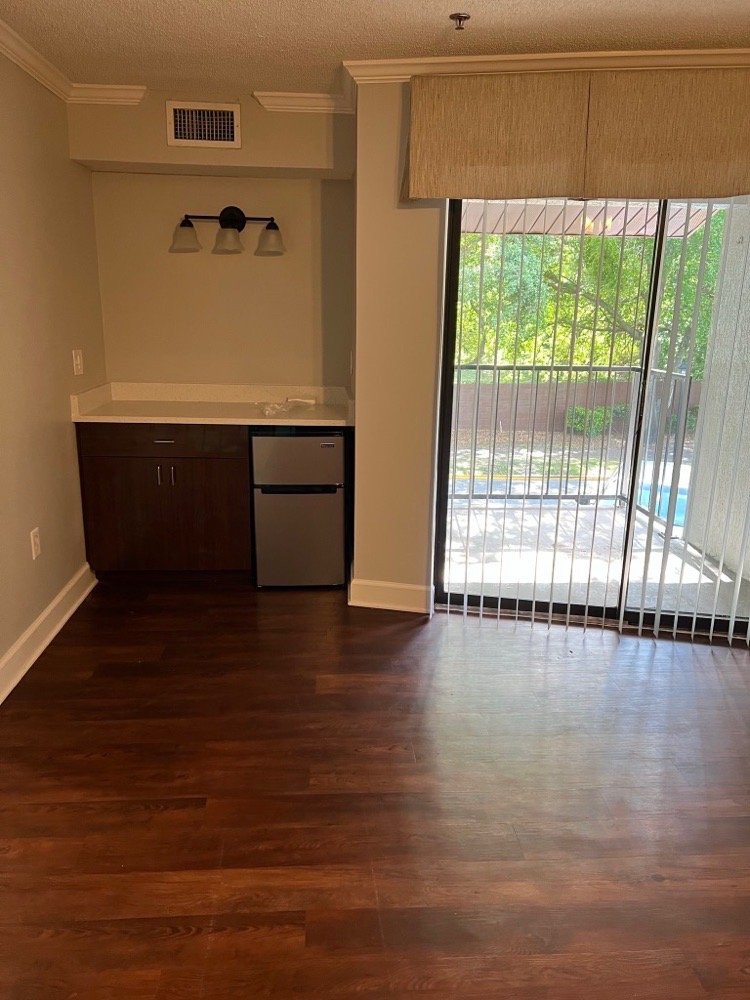An empty room with wooden flooring, featuring a small counter and mini fridge, and a sliding glass door with blinds leading to an outdoor area.