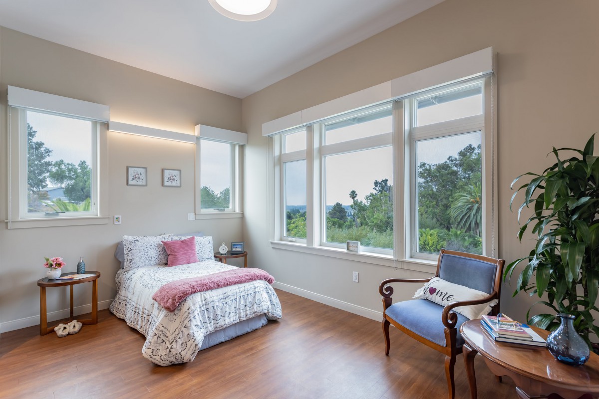 A cozy bedroom with a large window, allowing ample natural light. The room features a single bed, wooden flooring, and an elegant chair.