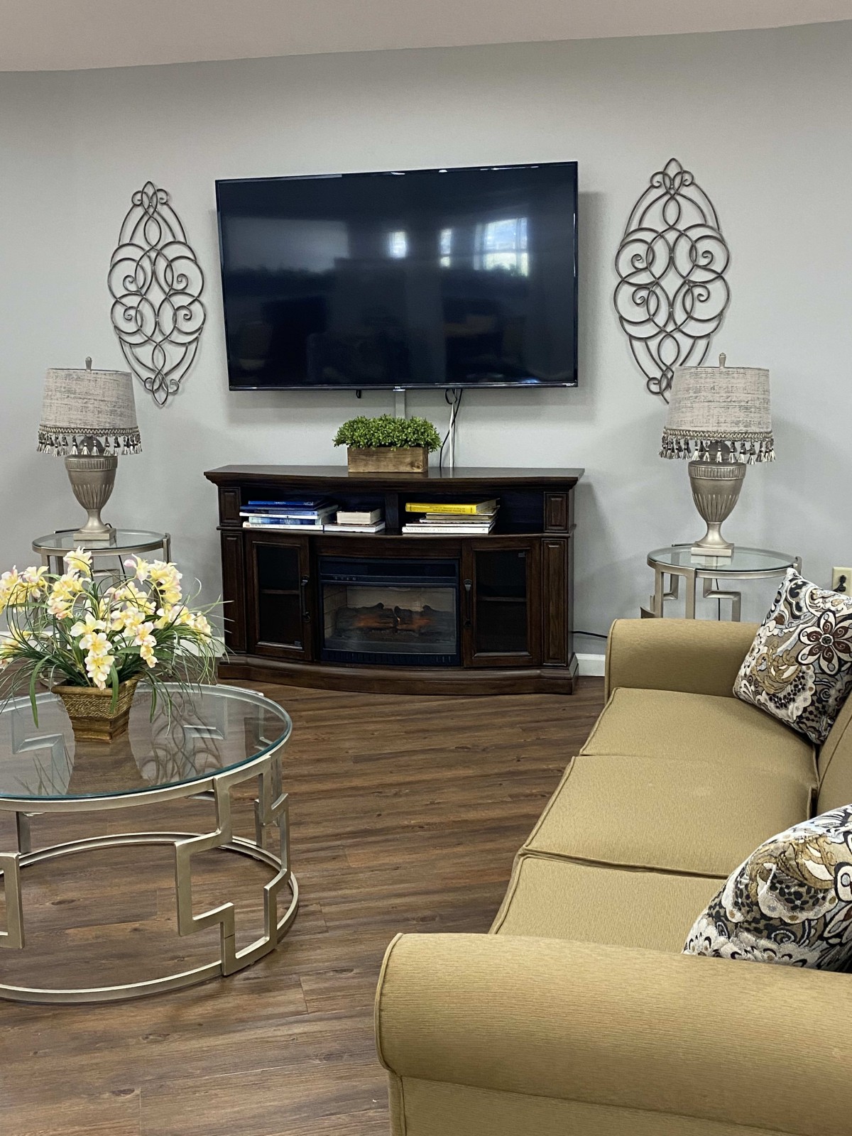 A cozy living room featuring a large sofa, a mounted television, decorative lamps, and a glass coffee table with flowers.
